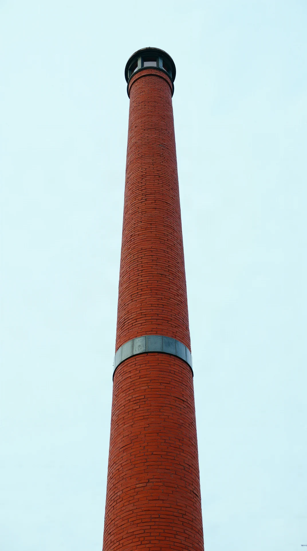A single tall red brick chimney stack with a metal flashing collar at its base, photographed straight on, chimney centred and filling the frame width, plain sof